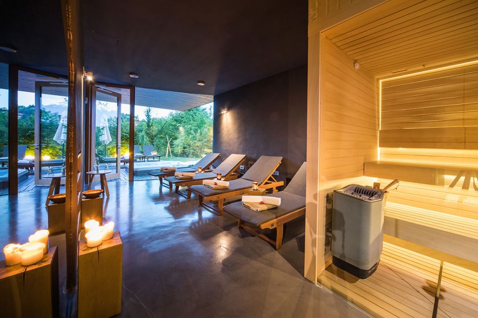 Wooden sauna for relaxation with glass walls and doors, illuminated by soft light. In front of the sauna are wooden chairs and candles